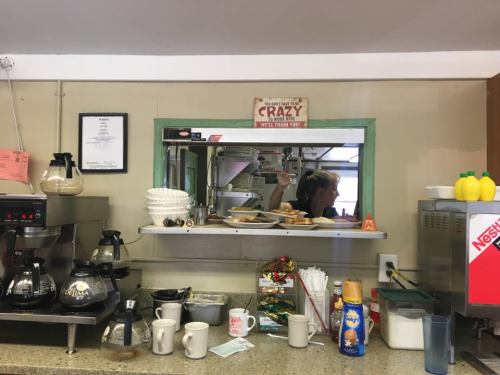A busy diner kitchen with coffee pots, plates of food, and a staff member preparing orders behind the counter.