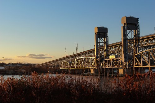 A large bridge spans a river at sunset, with tall support towers and vegetation in the foreground.