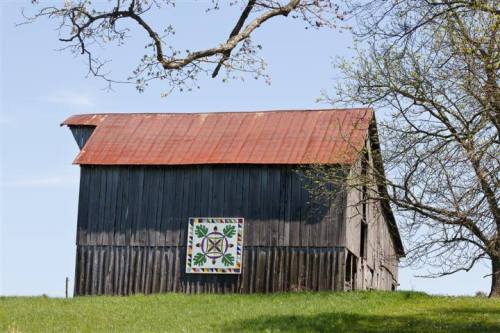 A rustic barn with a red roof, featuring a colorful quilt pattern on its side, set against a clear blue sky.