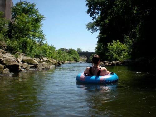 A person floats on a blue inner tube in a calm river, surrounded by greenery and a bridge in the background.