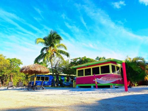 Colorful beach cottages with palm trees and a hammock under a blue sky. Sandy area with lounge chairs nearby.