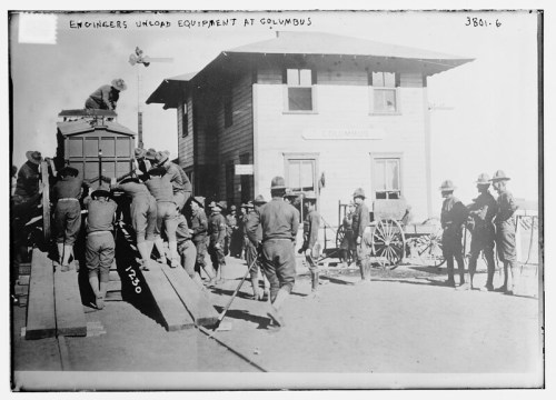 Engineers unload equipment at a train station in Columbus, with soldiers and a building in the background.