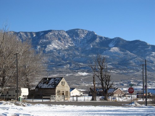 A snowy landscape featuring a rustic cabin, trees, and mountains under a clear blue sky. A stop sign is visible nearby.
