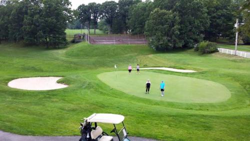 Two golfers on a green, with a sand trap nearby and a tennis court in the background, surrounded by lush grass.