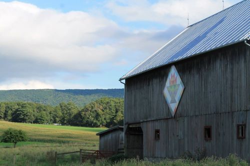 A rustic barn with a colorful quilt pattern on its side, set against a backdrop of rolling hills and blue skies.