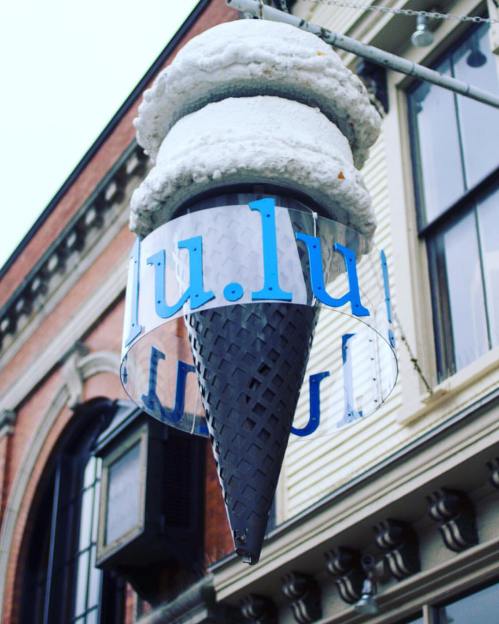 A large ice cream cone sign with a blue "lu.lu" logo hanging outside a building.