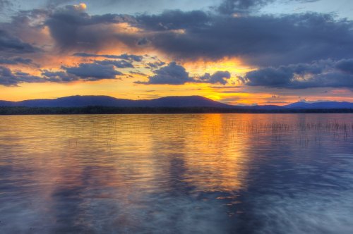 A serene lake at sunset, reflecting vibrant colors and clouds, with mountains in the background.
