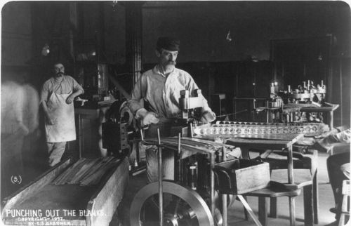 Two men working in a vintage workshop, one operating a machine while the other observes, surrounded by tools and materials.