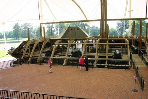 Visitors observe a historic wooden ship structure under a large canopy, with cannons visible inside.