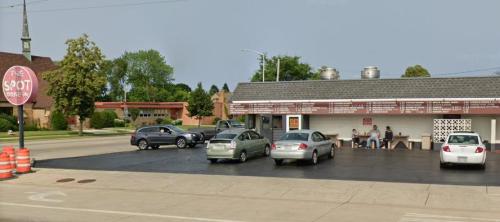 A small diner with outdoor seating, cars parked nearby, and trees in the background on a sunny day.