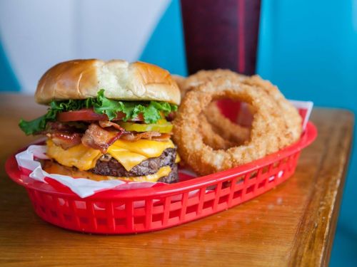 A juicy cheeseburger with bacon, lettuce, tomato, and pickles, served with crispy onion rings in a red basket.