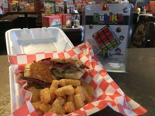 A sandwich with meat on a bun, served with tater tots, next to a Rubik's Cube in a casual dining setting.