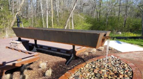 A large, rusted metal sculpture resembling a boat, set in a landscaped area with trees and stones.