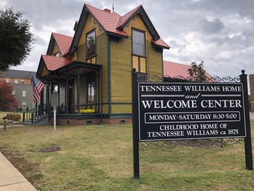 Historic yellow house with red roof, sign reading "Tennessee Williams Home and Welcome Center," surrounded by grass and trees.