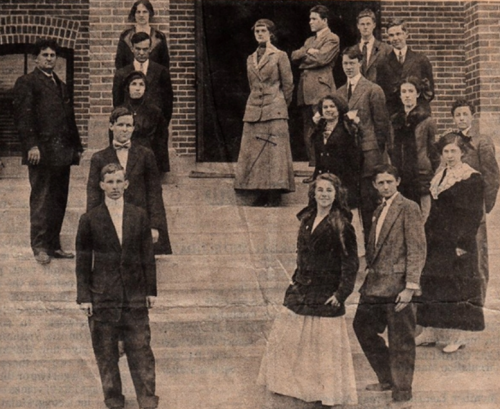 A historical group photo of men and women in early 20th-century attire, posed on steps of a brick building.