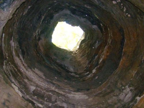 View looking up a cylindrical stone well, with light filtering through the opening at the top.