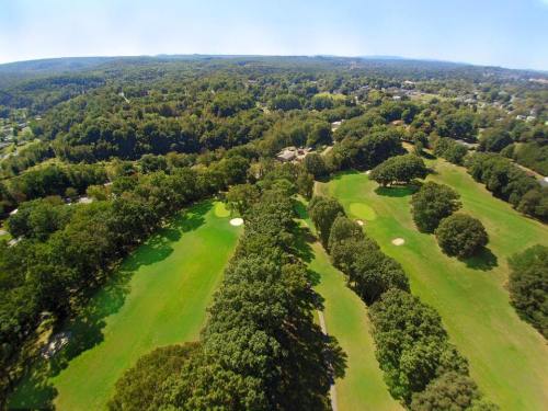 Aerial view of a lush green golf course surrounded by trees and hills under a clear blue sky.