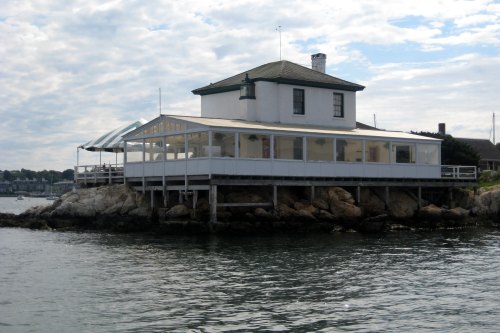 A white building on stilts over water, featuring a glass-enclosed patio and a green awning, surrounded by rocky shoreline.