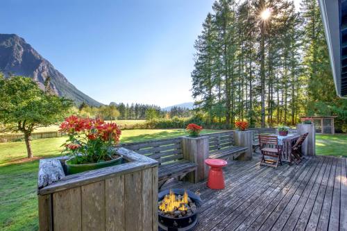A scenic deck with wooden seating, potted flowers, and a fire pit, surrounded by trees and mountains under a clear blue sky.