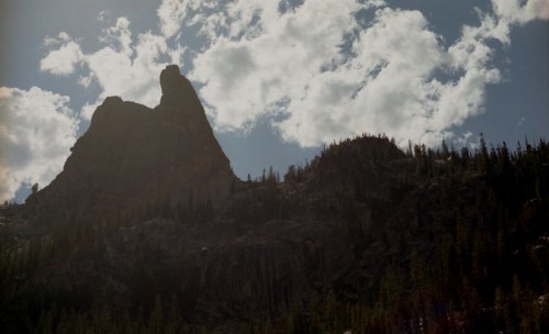 Silhouette of a rugged mountain peak against a cloudy sky, surrounded by dense forest.