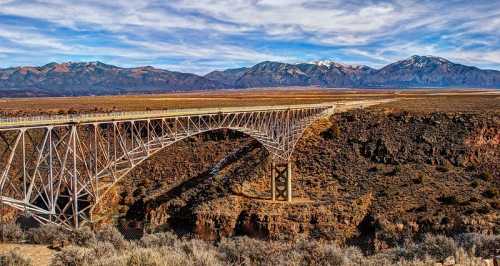 A large metal bridge spans a canyon, with mountains in the background and a clear blue sky above.