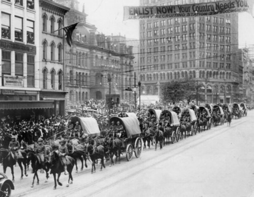 Historic black-and-white photo of a military parade with horse-drawn wagons and a crowd, featuring a recruitment banner.