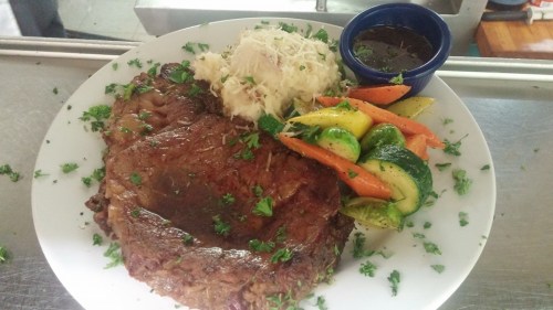 A plate featuring a large steak, mashed potatoes, mixed vegetables, and a small bowl of sauce, garnished with parsley.