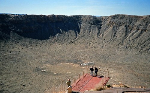 Two people stand on a viewing platform overlooking a large, dry meteor crater under a clear blue sky.