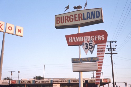 Sign for Burgerland featuring "Hamburgers 35" and various food options like tacos and pastrami, set against a clear sky.