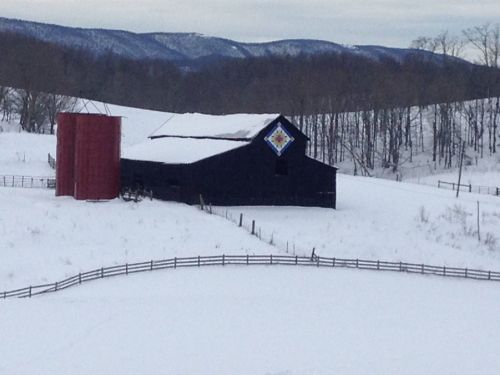 A black barn with a colorful quilt pattern stands in a snowy landscape, surrounded by trees and rolling hills.