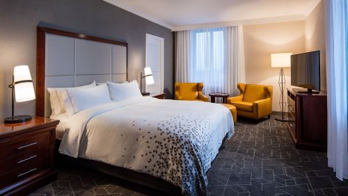 A modern hotel room featuring a king-sized bed, two yellow chairs, a TV, and large windows with sheer curtains.