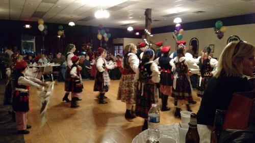 A group of dancers in traditional costumes perform in a festive hall decorated with balloons and tables.