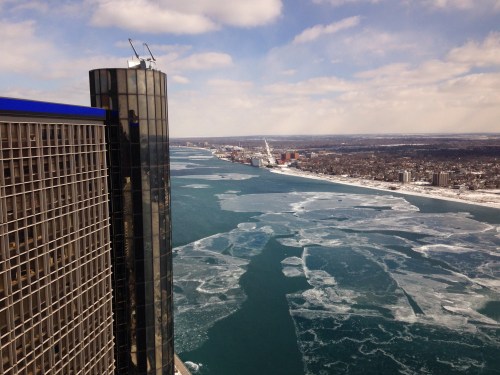 Aerial view of a city skyline with a river covered in ice and snow, under a partly cloudy sky.