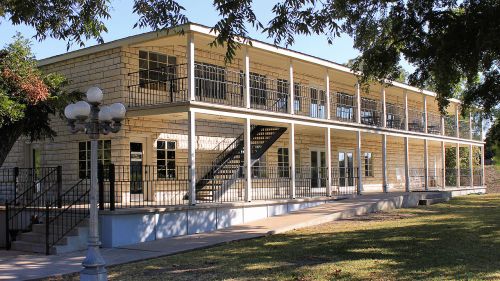 A two-story building with a light-colored exterior, balconies, and a staircase, surrounded by trees and grass.