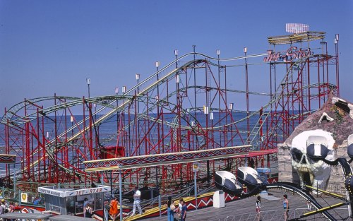 A colorful roller coaster named "The Star" on a boardwalk, with people enjoying the amusement park atmosphere.