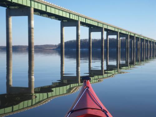 A red kayak glides on calm water, reflecting a green bridge above in a serene landscape.