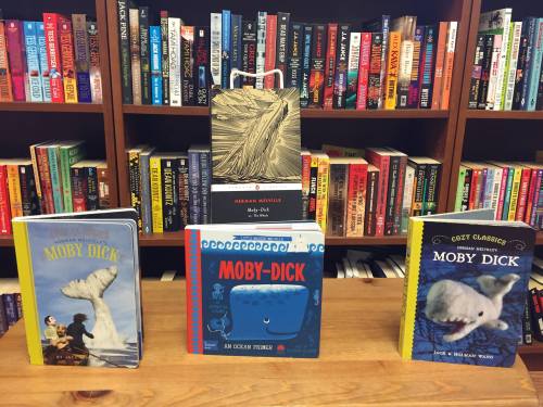 Three editions of "Moby Dick" displayed on a table, with a colorful bookshelf filled with books in the background.
