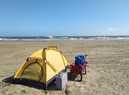 A yellow tent and a red cart on a sandy beach with ocean waves and a clear blue sky in the background.