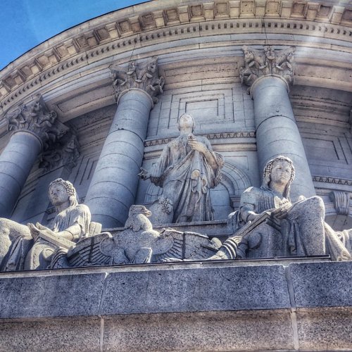 Close-up of ornate stone sculptures on a building facade, featuring figures and decorative elements against a blue sky.