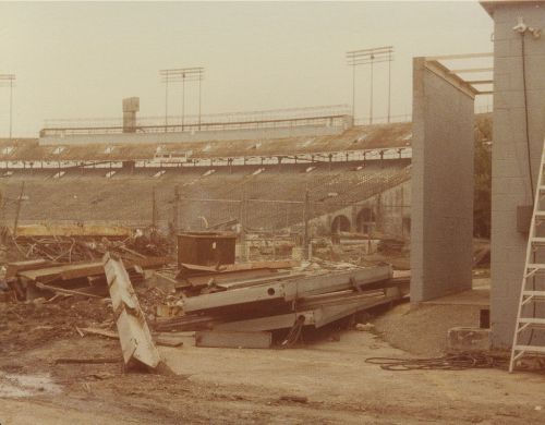 Construction site at a stadium, with debris and unfinished structures visible in a dusty environment.