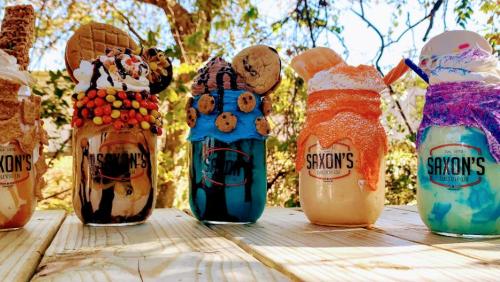 Five colorful milkshakes in jars, topped with cookies, candy, and whipped cream, displayed on a wooden table outdoors.
