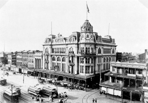 Historic black-and-white photo of a grand building with a dome, surrounded by streets and early 20th-century vehicles.