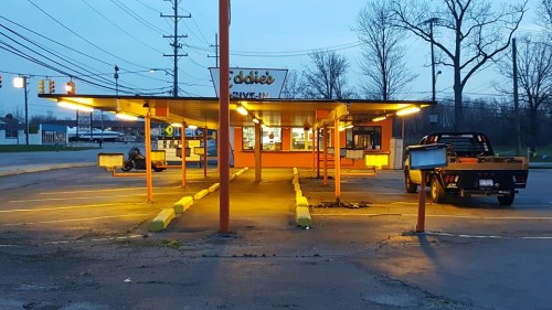 A retro drive-in restaurant with bright lights, empty parking spaces, and a pickup truck parked nearby at dusk.