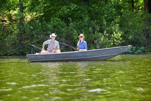 Two people fishing from a small boat on a calm lake, surrounded by lush greenery.