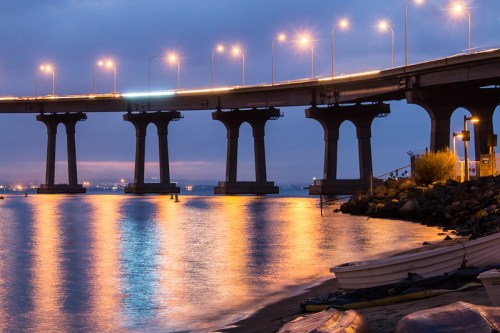 A bridge illuminated at dusk, reflecting lights on calm water, with boats along the shore and a cloudy sky above.