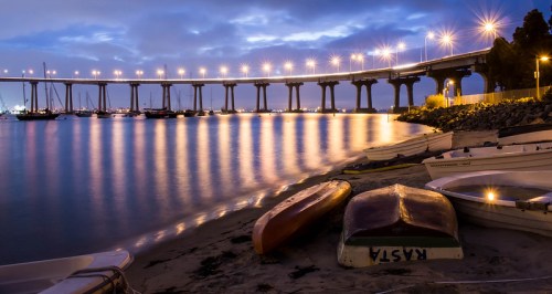 A serene night scene of a bridge illuminated by lights, reflecting on calm water with boats on the sandy shore.