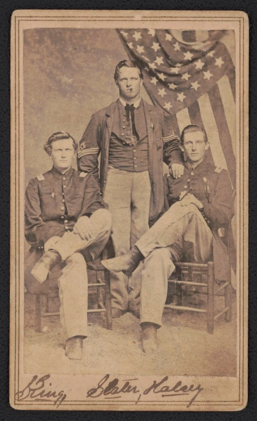 Three men in Civil War uniforms pose for a portrait, with an American flag in the background.