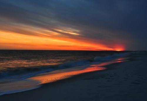 A serene beach at sunset, with vibrant orange and purple hues reflecting on the water and gentle waves lapping the shore.