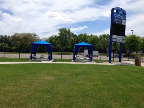 Two blue canopies set up on a grassy area, with a sign and trees in the background under a partly cloudy sky.