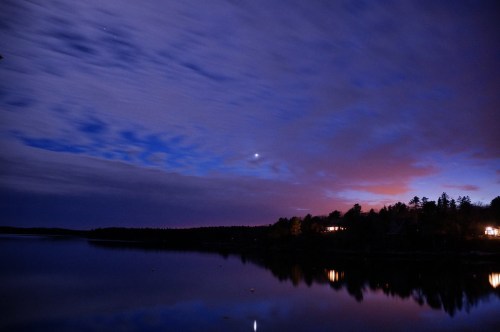 A serene lake at dusk, reflecting colorful clouds and a crescent moon, with trees and distant lights along the shore.
